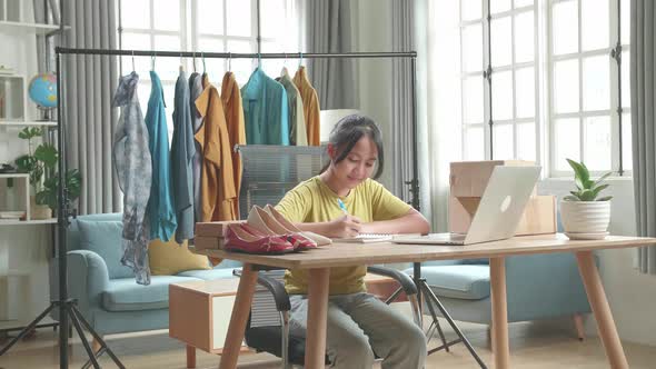 Asian Young Girl Online Seller Looking At Computer Screen And Writing While Selling Clothes At Home