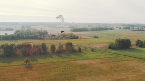 Aerial View of a Paramotor Gliding Through the Air Over Green Fields