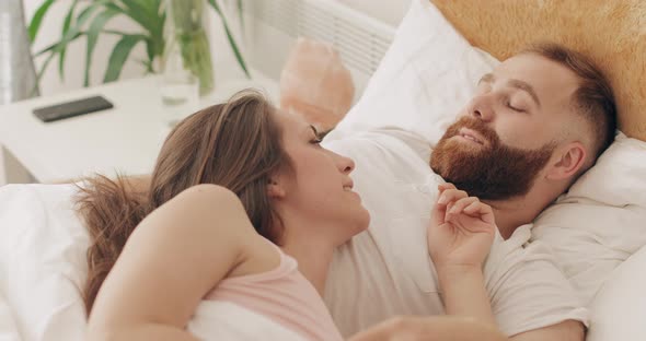 Cheerful Couple Lying on Bed in Early Morning, Discussing Plans for Day and Laughing. Beautiful