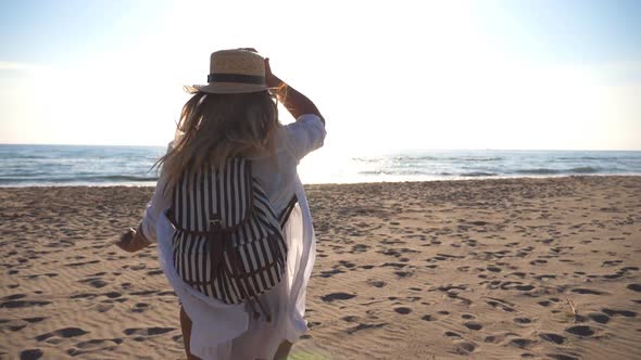 Happy Girl in Bikini and Shirt with Backpack Running on the Beach to the Sea