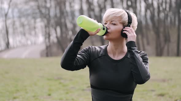 Athletic Woman in a Mask Is Listening To Music in Earphones and Drinking Water