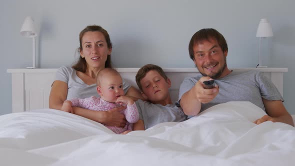 Happy Young European Family with Two Kids Gather on Couch to Watch TV in the Badroom Smiling