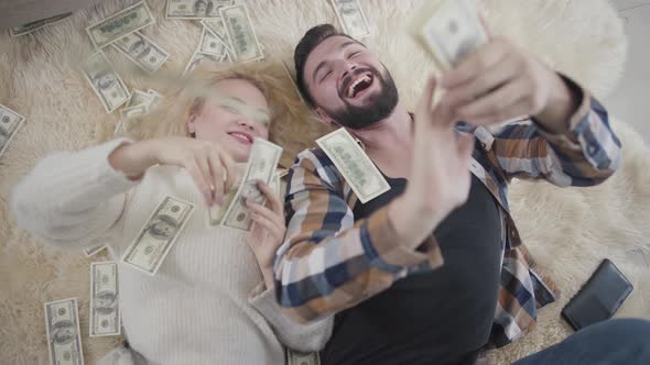 Top View of Wealthy Caucasian Husband and Wife Lying on Soft Carpet and Scattering Money. Happy
