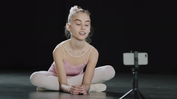 Caucasian Girl Child Teen Ballerina Gymnast Sitting on Floor in Dance Hall Waving Hello to Mobile