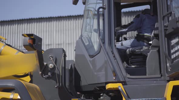 Side View of Caucasian Driver Sitting in Cabin of Yellow Loader. Young Employee in Uniform Inside