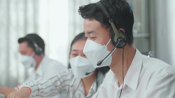 Man And A Woman Of Three Asian Call Centre Agents In Headsets And Masks Discussing About Work