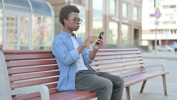 Young African Man Celebrating Online Success on Smartphone While Sitting Outdoor on Bench