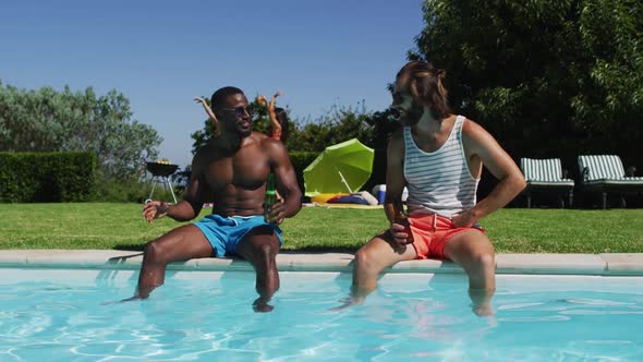 Two male diverse friends holding beer talking to each other while sitting by the pool