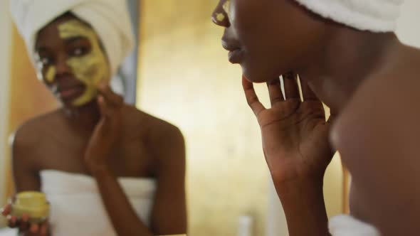 African american woman with towel watching in mirror and using cream on her face in bathroom