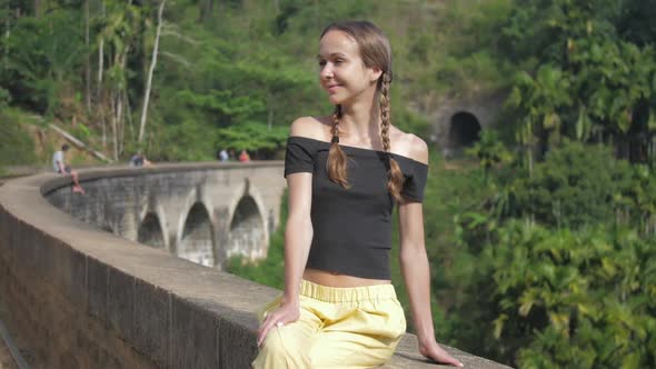 Girl Sits on Historical Bridge Smiling Against Wild Forest