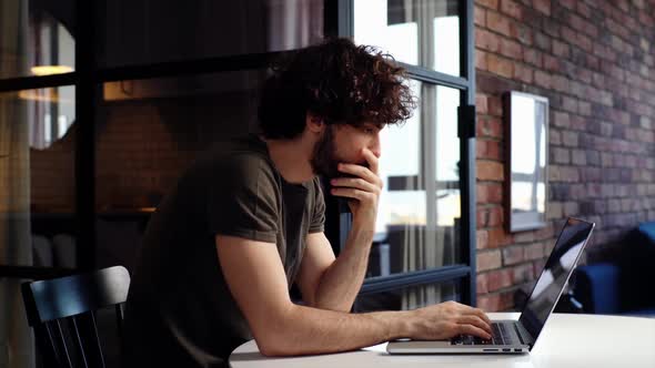 Side View of Pensive Young Man Programming on Laptop Sitting at Desk at Home Office