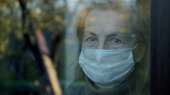 Elderly Woman in a Window Wearing a Protective Mask Looks Into the Camera