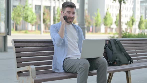 Angry Young Man with Laptop Talking on Phone While Sitting on Bench