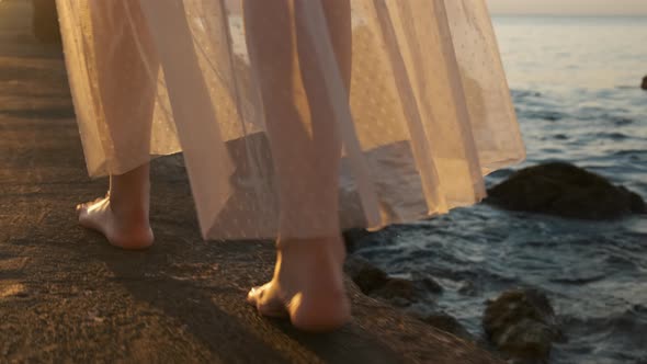 Woman in White Fluttering Dress Walking Along the Stone Coast Near the Sea at Sunlight