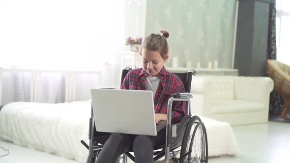 Portrait of a Teenage Girl in a Wheelchair Uses a Laptop at Home in the Bedroom