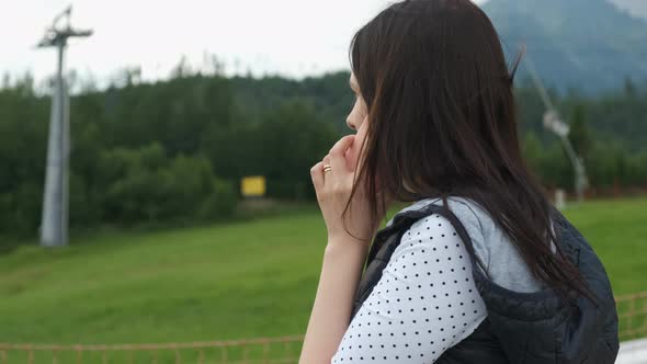 Woman in a Warm Vest Talking Smartphone Standing on the Background of Mountains and Moving Ski Lifts