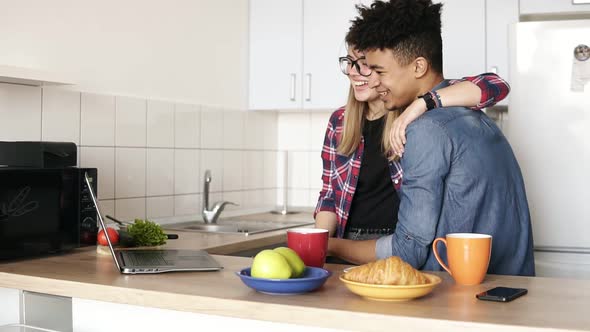 Cute Couple of Two Young People Cuddling and Using Laptop Surfing the Web in the Kitchen