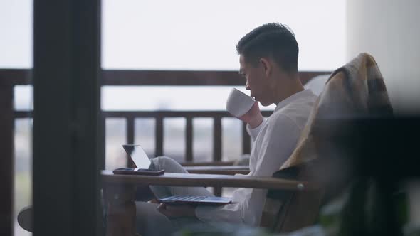 Nervous Troubled Young Asian Man Drinking Coffee Typing on Laptop Keyboard Sitting on Armchair on