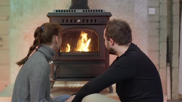 Young Loving Couple Sitting By Fireplace Watching Wood Burning Resting at Home