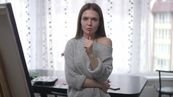 Portrait of Exhausted Young Woman with Painting Brush Looking at Camera Standing Indoors at Easel