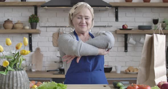 Portrait of Happy Senior Caucasian Woman Posing in Kitchen. Middle-aged Lady in Apron Crossing Hands