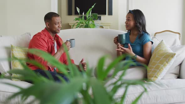 Happy biracial couple sitting on sofa with coffee and talking