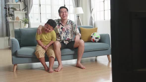 Happy Asian Father And His Son Are Watching Tv While Sitting On A Couch At Home