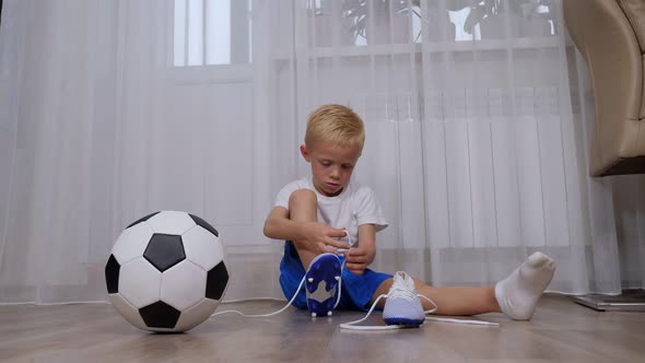 A Little Boy Sits on the Floor and Learns To Tie the Laces on Football Boots