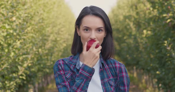 Young Woman Biting Fresh Natural Apple Standing in Own Fruit Garden and Smiling