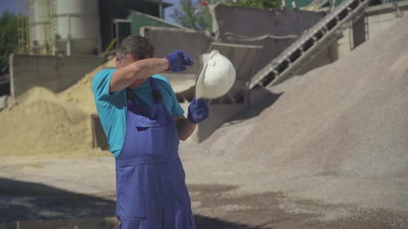 Portrait of Exhausted Factory Worker Taking Off Safety Helmet, Wiping Forehead with Hand, and