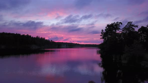 Aerial backward drone shot, bypassing a man sitting at a rocky shore, of a lake, a purple sky, a col