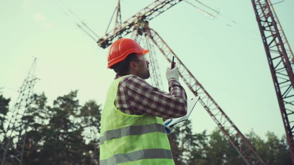 Caucasian Engineer in Protective Uniform and Orange Hard Hat Holds Walkietalkie and Clipboard on