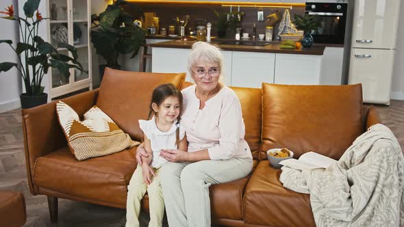 Granny and Granddaughter are Smiling and Hugging Sitting on Sofa with Cookies in Bowl Studio