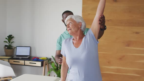 Video of happy caucasian senior woman exercising with african american male physiotherapist