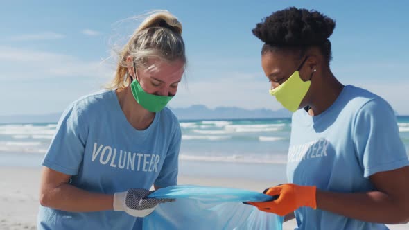 Two diverse women wearing volunteer t shirts and face masks picking up rubbish from beach