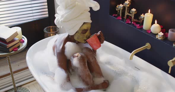 African american woman with towel and mask taking bath and drinking coffee in bathroom