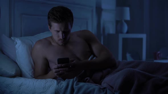 Handsome Male Lying in Bed, Reserving Disappointing Text Message on Smartphone