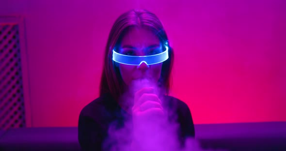 Young Woman Soars Vape in Neon Light in Cyber Glasses