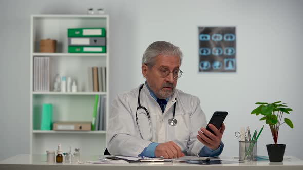 Aged Male Doctor Fills in Medical History and Advises Patient on Video Call Using Mobile Phone About