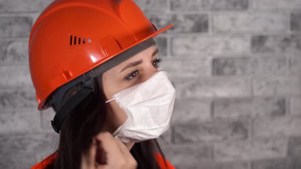 Female Construction Worker in Overalls Removing Medical Mask From Face on Background of Gray Brick