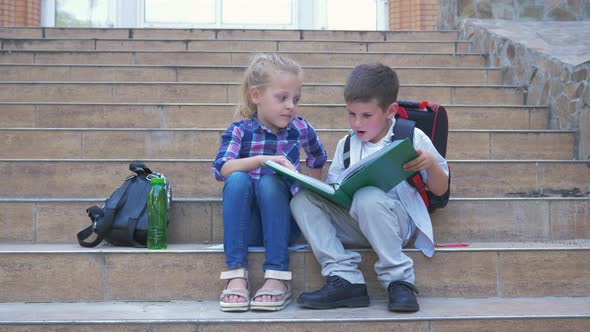 Pupils Kids with Backpacks Reading and Leafing Through a Book Sitting on Steps of the School