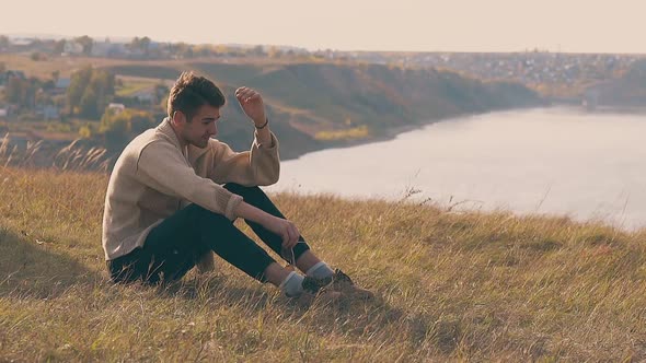 Frustrated Guy in Jeans and Pullover Sits on River Bank