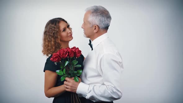 A Happy Dancing Middleaged Couple in Love with a Bouquet of Scarlet Roses in Their Hands on a White