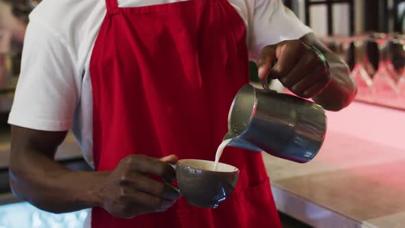 Midsection of african american barista pouring milk from jar into coffee cup in cafe