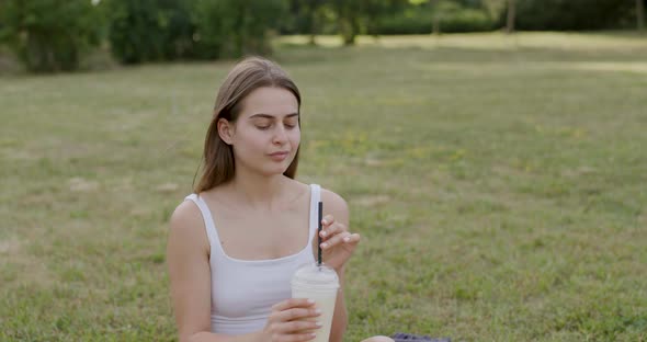 a Girl Filmed Up Close Dressed in White Drinking a White Drink in the Park