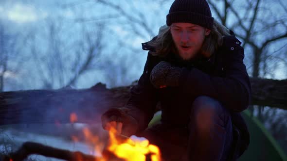Portrait of Caucasian Unshaved Guy Wearing Hat Smiling While Sitting Near Bonfire in Forest and