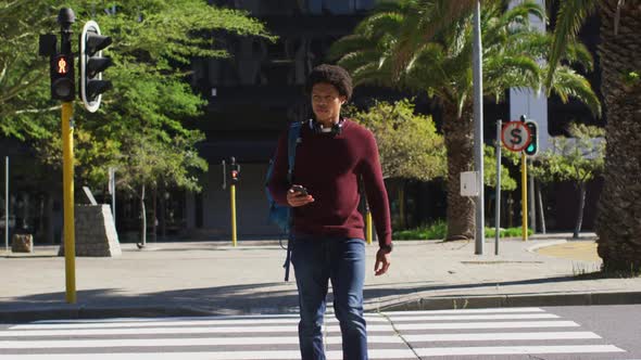African american man in city, using smartphone, wearing headphones and backpack crossing street