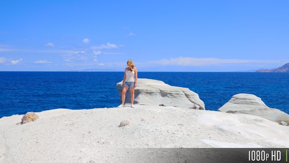 Moving toward woman on water's edge with steep cliffs and ocean behind her looking at camera