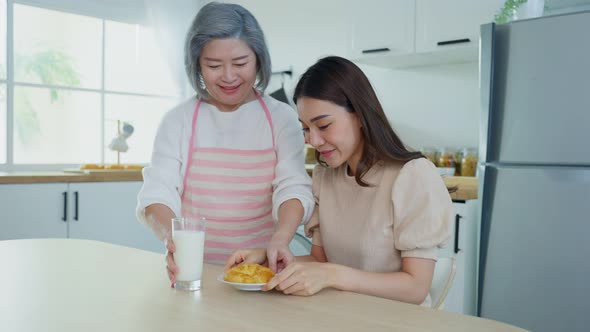 Asian loving senior elderly mother serve foods by handing croissant and milk to daughter in kitchen.