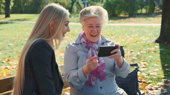 Beautiful Blonde Girl with Elder Woman Sitting on Bench and Looking Something on Smartphone